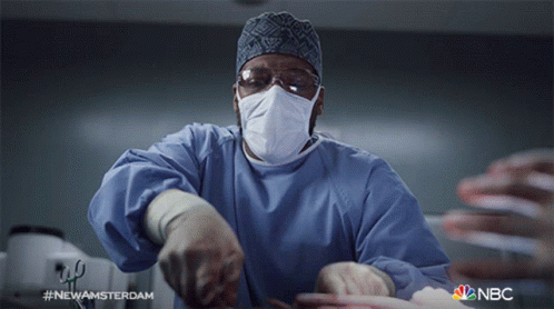 O arquivo gif mostra um cirurgião negro com um bisturi em uma sala de cirurgia. Ele está ofegante e olha para cima com preocupação.