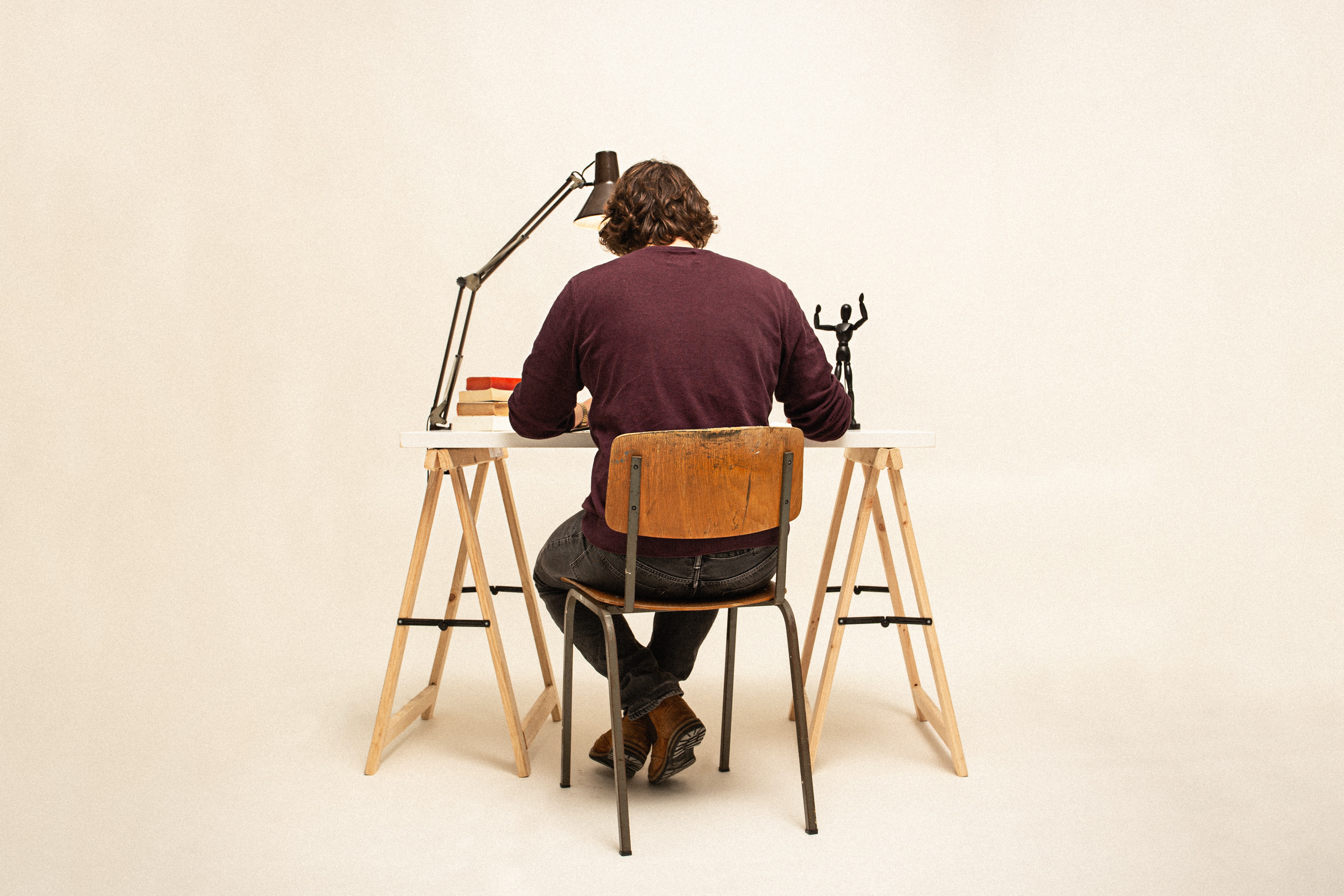 Imagem de um homem sentado em uma cadeira, de costas para nós, e com a cabeça baixa para uma mesa que possui uma lâmpada, um boneco, cadernos e mais alguns objetos.