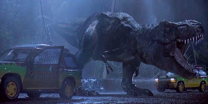 Tiranossauro Rex rugindo entre dois carros em uma cena noturna com chuva e uma luz artificial forte vinda de cima.