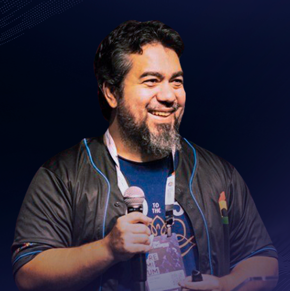 Imagem de Luciano Martins, Developer Advocate, Google IA. Luciano está com uma camiseta azul e uma jaqueta preta, um microfone está sendo segurado na sua mão direita e o fundo é um gradiente azul.