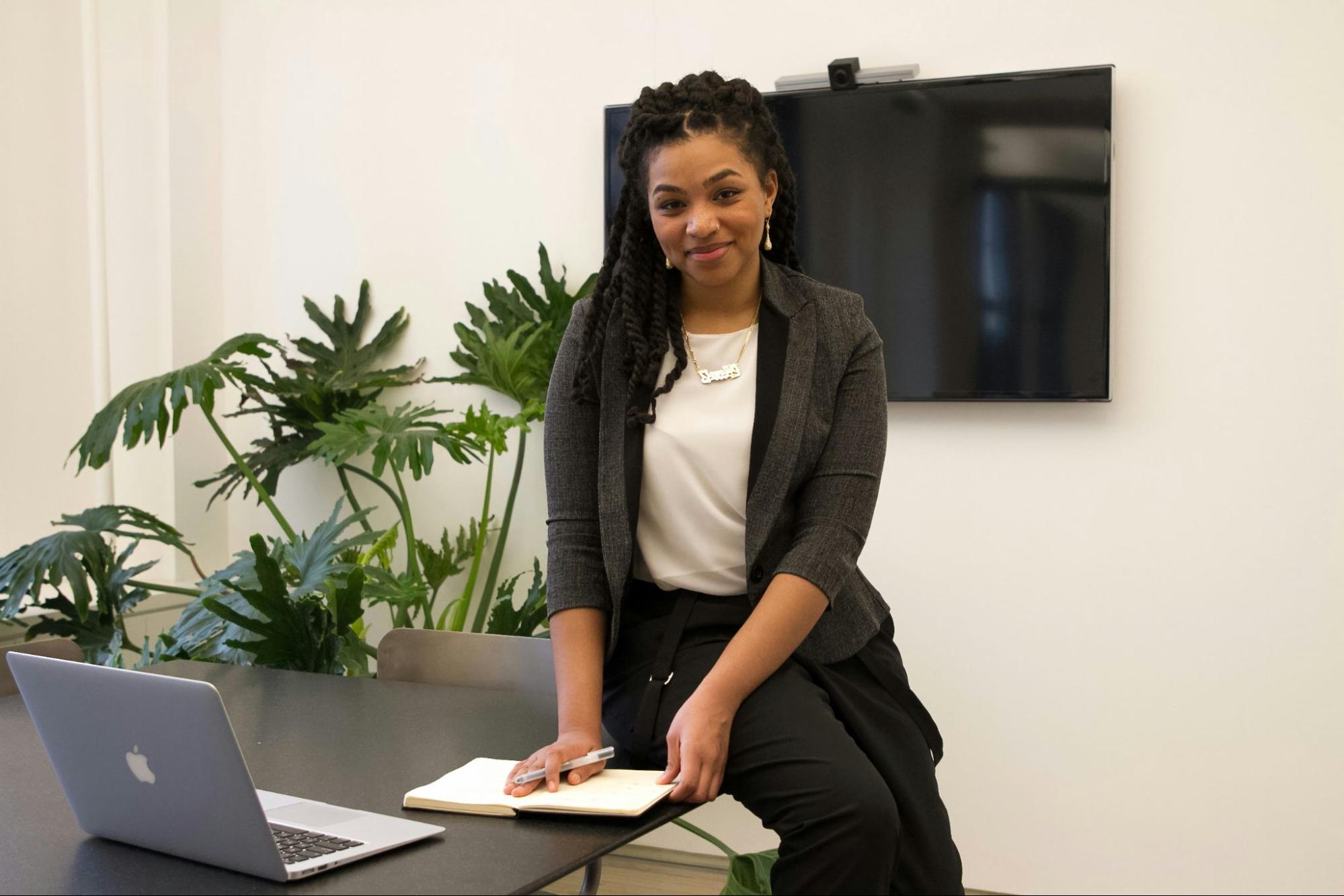 Mulher sorrindo, sentada na beira de uma mesa com um laptop e bloco de notas, em um escritório com plantas ao fundo.