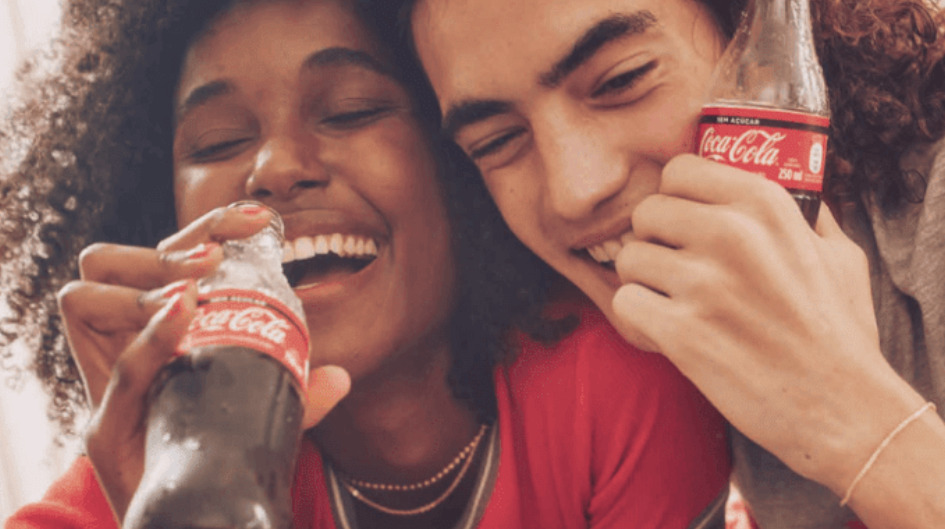 A imagem é de uma campanha da Coca-Cola. Aparece duas pessoas abraçadas tomando refrigerante.