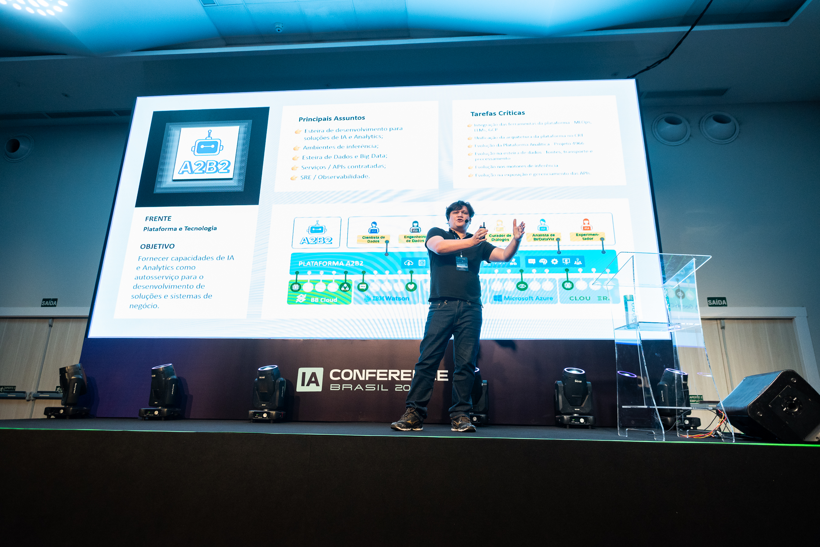 Na fotografia, Fábio aparece no palco com calça e camiseta pretos e, ao fundo, um slide de apresentação