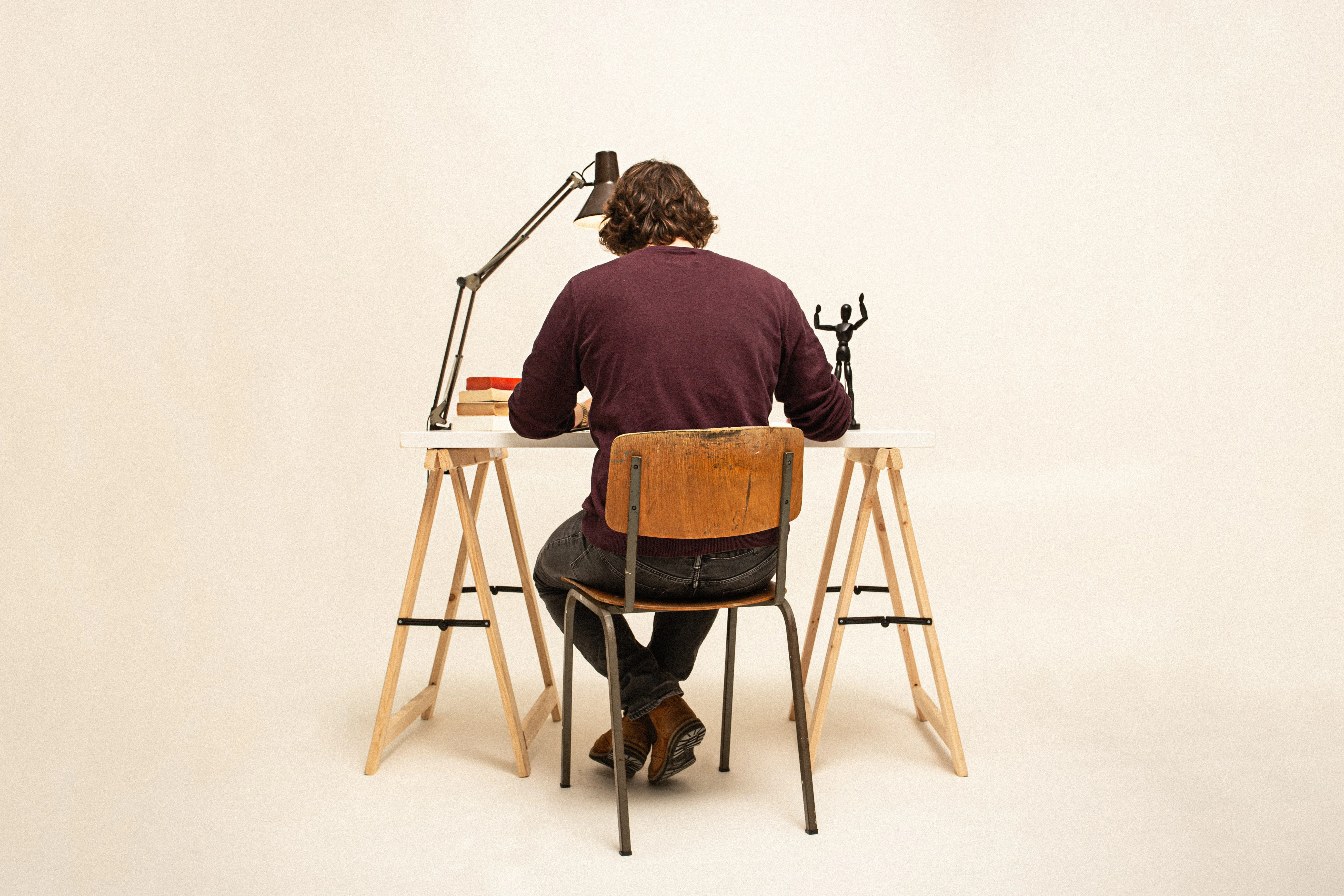 Imagem de um homem sentado em uma cadeira, de costas para nós, com a cabeça levemente abaixada na direção da mesa à frente dele, a qual possui uma lâmpada, um boneco e cadernos fechados de diferentes cores.