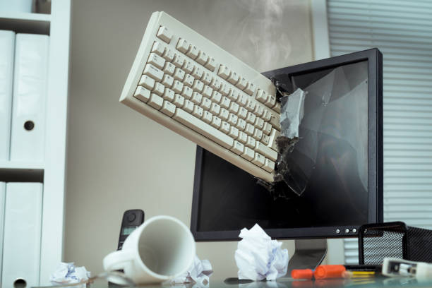 A imagem mostra um teclado branco enfiado no meio da tela de um computador preta. O centro da tela está quebrada pelo impacto do teclado, como se a dividisse fisicamente em duas partes e há algumas folhas de papel sulfite brancas, uma caneca branca vazia derrubada em cima da mesa e algumas canetas coloridas.