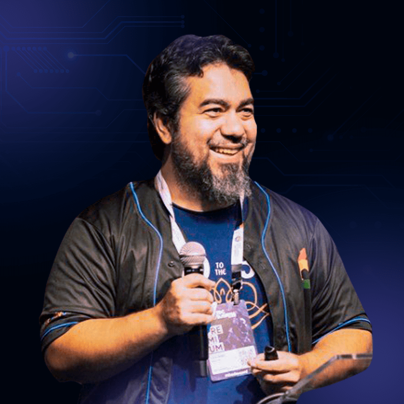 Imagem de Luciano Martins, Developer Advocate, Google IA. Luciano está com uma camiseta azul e uma jaqueta preta, um microfone está sendo segurado na sua mão direita e o fundo é roxo com elementos gráficos que remetem à tecnologia.