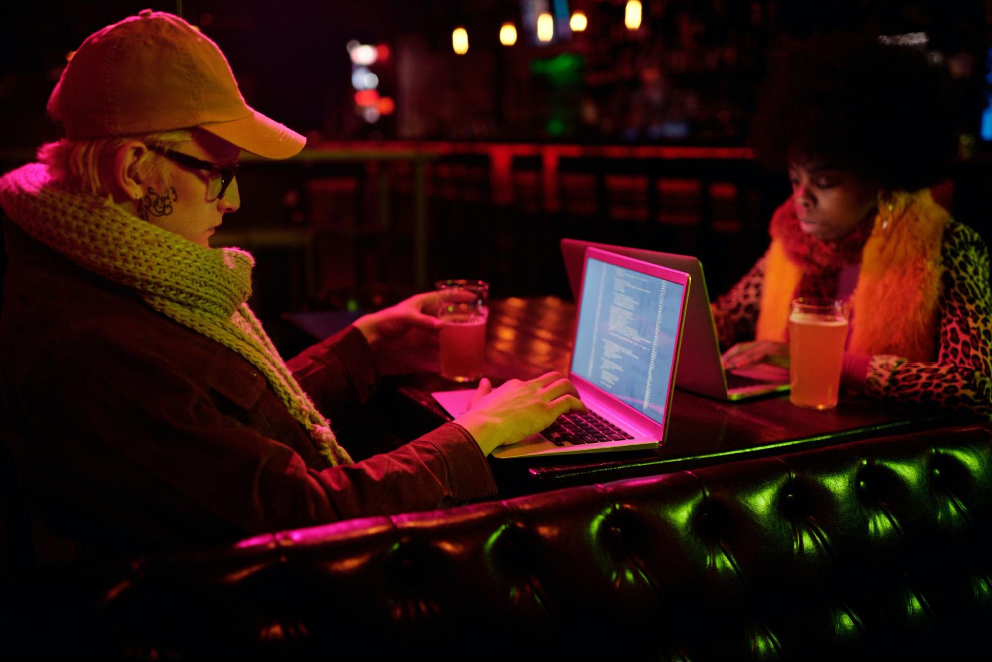 Duas pessoas sentadas em uma mesa de um bar, usando o computador para escrever códigos de programação.