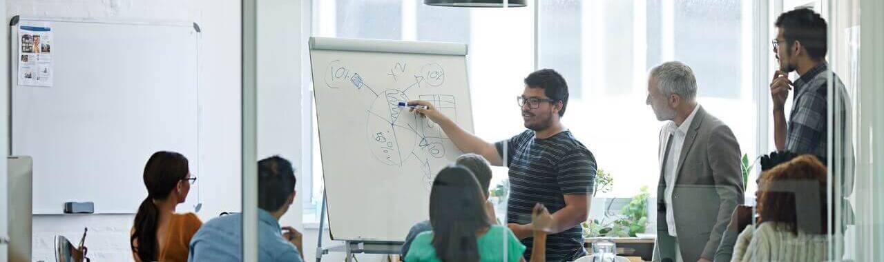 A imagem mostra um grupo de pessoas em uma reunião, com um homem apresentando ideias em um quadro branco, enquanto os demais participantes escutam e discutem, representando a semana de 4 dias